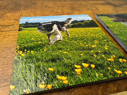 A square printed coaster or tile featuring a black-and-white dog running through a green field of yellow flowers, placed on a wooden tabletop. A second matching printed item is partially visible at the right edge.