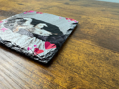 A square slate coaster with a printed design of a sleeping dog on a gray patterned background with pink hearts, resting on a wooden table.